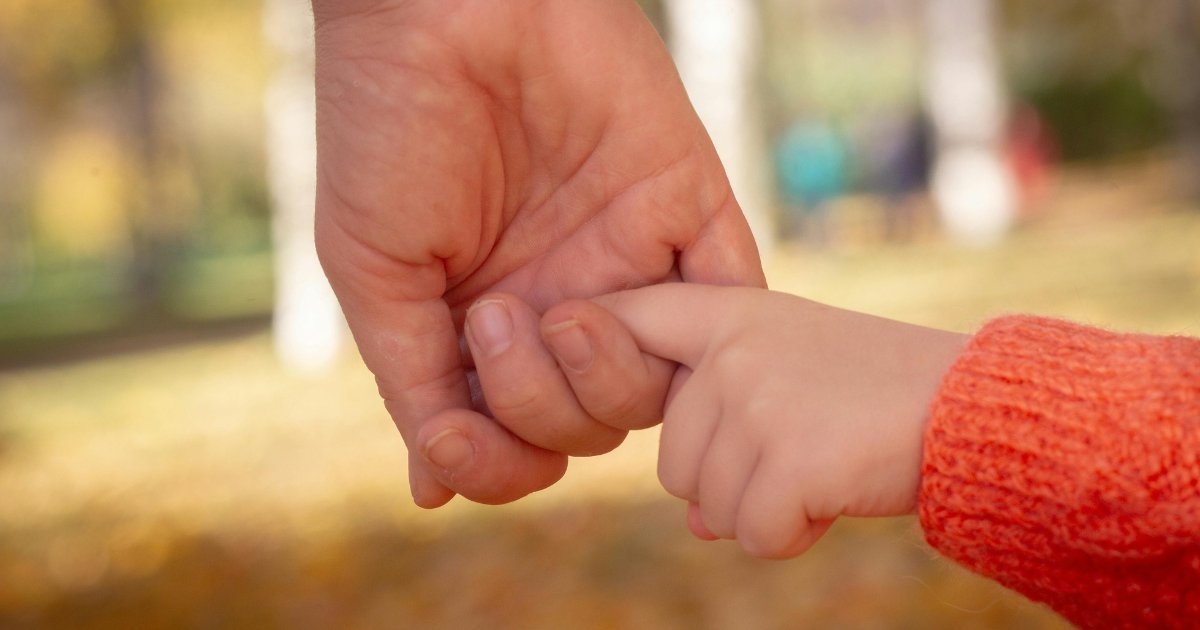 Une main d'adulte qui tient la petite main d'un enfant, dans un cadre extérieur baigné de lumière chaude